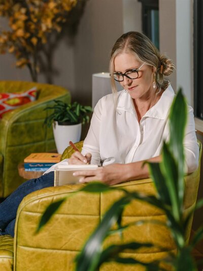 Phillipa Leseberg, with glasses on, sits on a green chair, authoring her latest book, "His Name is Eric," in her notebook. A plant and books are in the background.