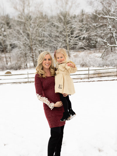Mom pregnant with baby holding her little girl during a snowy maternity shoot outdoors