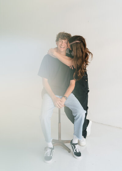 Woman kissing man with her arms around him while he sits on a stool in front of a white studio background