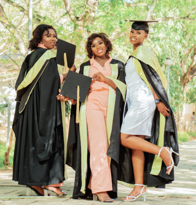 3 women wearing dresses, heels, and graduation caps and gowns posing on campus in front of trees