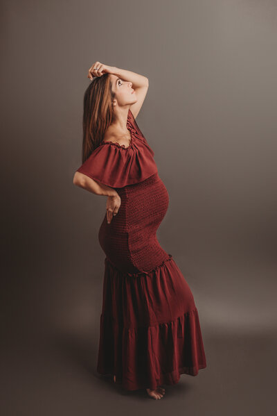 Pregnancy portrait of 37 week pregnant lady wearing maroon fitted maternity gown with hand on head and other hand on back, facing to the side on dark gray backdrop