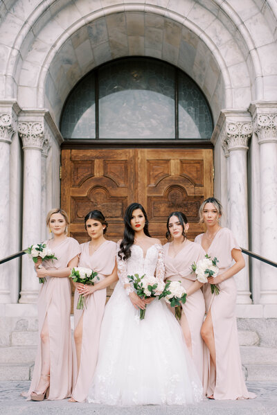 dark haired stunning bride stand outside of a Florida Spanish cathedral  with her bridesmaids who are in all light pink holding white rose bouquets, best wedding photographer Tampa Florida