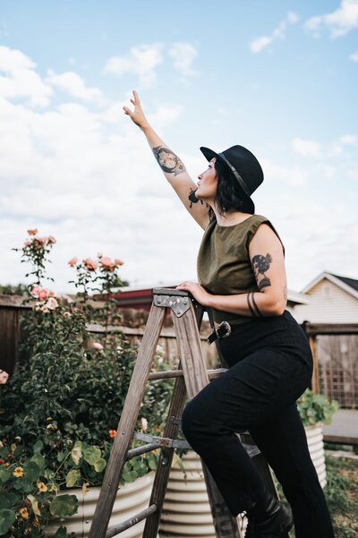 Woman with tattoos wears a black hat and dark green sleeveless top standing outside in a garden on a ladder reaching toward the sky.