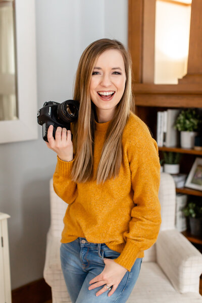 Abby is smiling at the camera, wearing a mustard yellow sweater and holding a camera