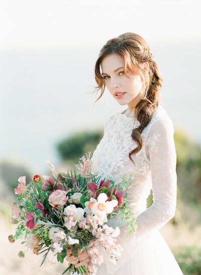 Kohnur romantic bride with soft natural makeup and braided side ponytail, holding a bouquet on a bright outdoor hilltop location.