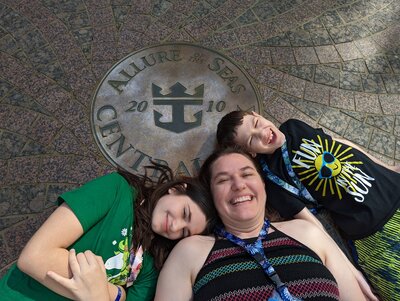 A mom and her two kids lie on the floor, grinning up at the camera. Their heads rest along the edges of a circular seal in the ground reading Allure of the Seas, Central Park, 2010.