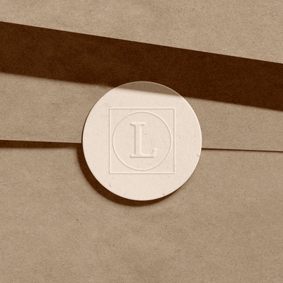 A close-up of a round, textured sticker with the letter "L" embossed in the center. The sticker is placed on a piece of brown paper, creating a minimalist and elegant look.