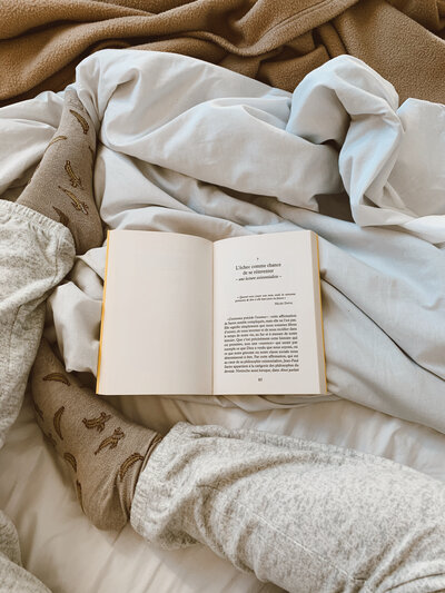 Cozy scene with an open book on a bed, flanked by gray pajama-clad legs and warm socks. Soft, crumpled blankets add a relaxed, comforting vibe.