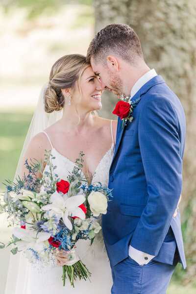 A groom and bride stand forehead to forehead as she holds her bouquet of red, white and blue flowers.