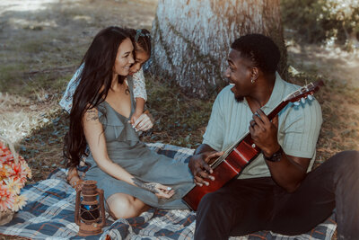A sweet family moment at Prospect Park in Redlands, with dad playing guitar during a cozy picnic while mom and daughter share laughter and cuddles under the trees