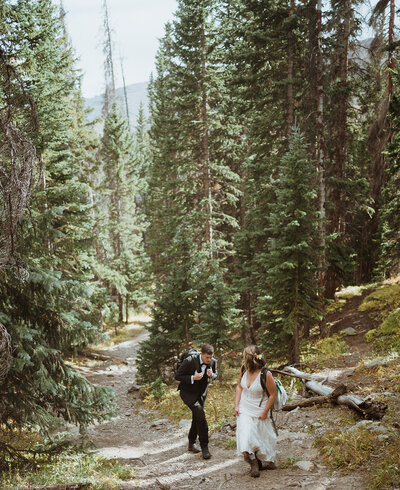 bride and groom are hiking up the trail with trees surrounding them. the bride is looking back at the groom as he is holding onto his backpack. the bride has flowers in her hair and is holding her dress up to reveal that she is wearing hiking boots.