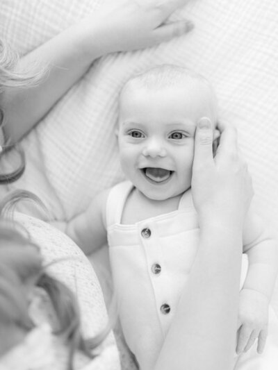 A baby boy is laying on a bed and smiling as he looks up at his mother.