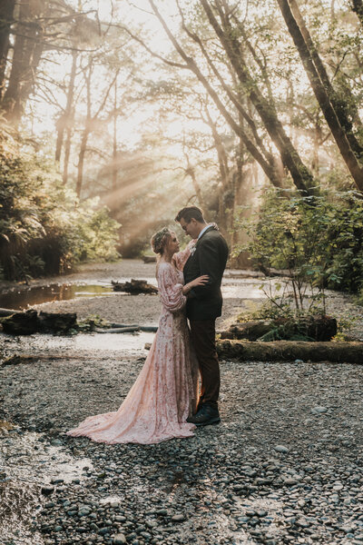 Bride wearing pink wedding dress and flowers in hair faces groom wearing green suit jacket looking into each other's eyes in Fern Canyon surrounded by light rays and trees taken by California elopement photographer Kasey Mantiply
