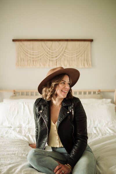 woman sitting on bed with brown hat and leather jacket smiling during photo shoot