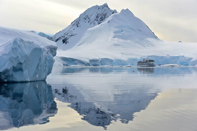Snow-covered mountain reflected in calm Antarctic waters with a small cruise ship in the distance.