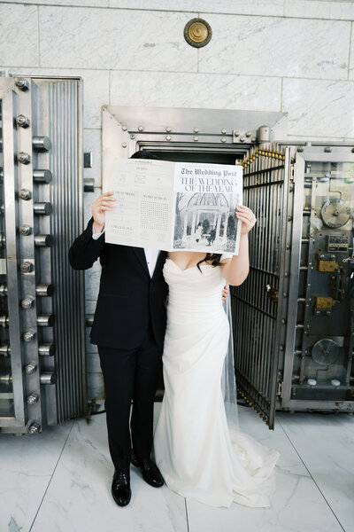 Bride and groom holding up a wedding newspaper in front of their faces
