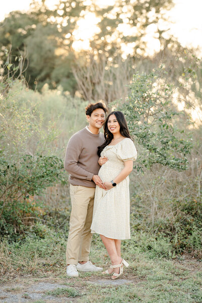 The picture captures a serene and intimate moment between a young couple during a maternity session in the dry Texas winter. The woman, dressed in an off-white dress that perfectly showcases her baby bump, stands with her partner, who is wearing a brown sweater and khaki pants.  The couple's pose is affectionate, with the man's arm wrapped around his partner's shoulder and his other hand resting gently on her belly. They stand in a dry, grassy field, with a backdrop of a dry forest at sunset, creating a warm and natural setting.  The photographer, Bri Sullivan, has captured the scene in a light and airy style, highlighting the couple's connection and the beauty of the expectant mother's form. The soft, muted tones of the image add to the serene and intimate atmosphere, drawing the viewer's attention to the couple's tender embrace and the anticipation of their growing family.  The dry, winter landscape provides a unique and striking contrast to the couple's warm and loving embrace, creating a sense of timelessness and a connection to the natural world. Overall, the picture is a beautiful and heartwarming representation of the joy and wonder of pregnancy, captured with skill and artistry by the talented photographer.