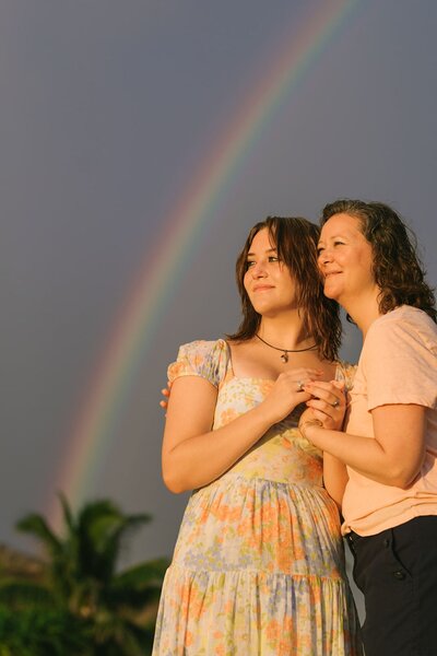 A mother holds her older daughter with a rainbow in the background.