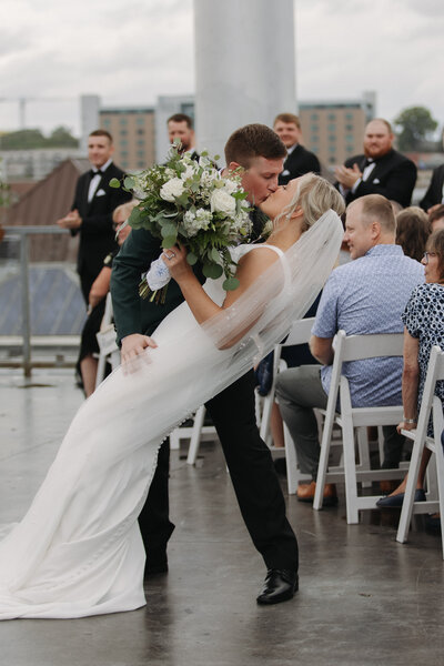 The bride playfully runs away as the groom chases her. She is dressed in a mid length satin white dress and has a short veil flowing behind her.