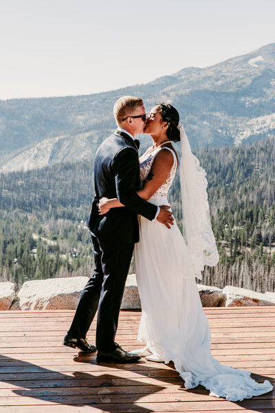 A bride and groom share a romantic kiss on a wooden platform overlooking a breathtaking mountainous landscape, bathed in bright sunlight, near Lake Tahoe