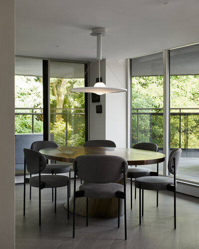 A spacious dining room featuring a large, round wood dining table surrounded by soft grey chairs, a diffused globe pendant light, and tall windows