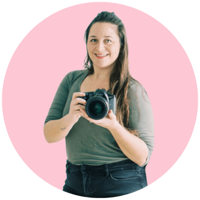 Sara, wearing a green shirt and black pants, holds her camera in her hands and smiles.