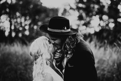 brudgum med hatt och långt hår kysser brud med bohemisk klänning under bröllopsfotografering