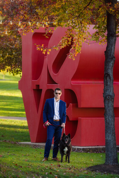 Groom standing with dog in front of large love statue