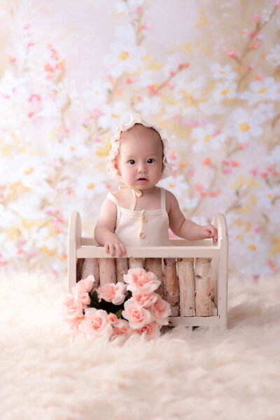A baby in a cream outfit and bonnet sits in a wooden crate adorned with pink roses. The background features soft floral patterns, creating a gentle, dreamy atmosphere.