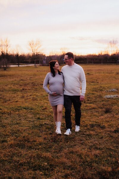 Mann und Frau mit Babybauch laufen während eines Sonnenuntergangs auf einer Wiese