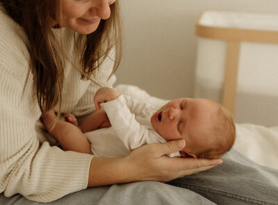 A woman in a cream color sweater poses in her living room in Colchester, Vermont. She sits smiling while holding her sleeping newborn baby. A perfect setting for capturing cozy moments by a New England Portrait Photographer.