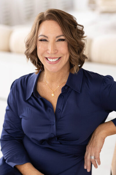 A smiling woman with short, wavy brown hair wearing a navy blue blouse, seated indoors with a relaxed posture.