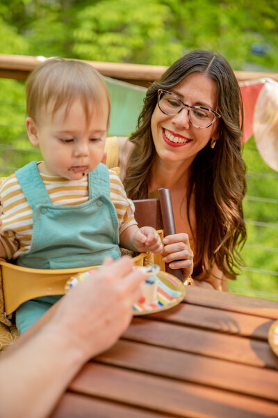 heather of sweet heather anne smiles over shoulder of toddler son as he enjoys birthday cake outdoors