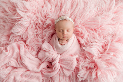 "Newborn baby girl wrapped in soft pink fabric with a delicate floral headband, posed peacefully on a fluffy pink backdrop.