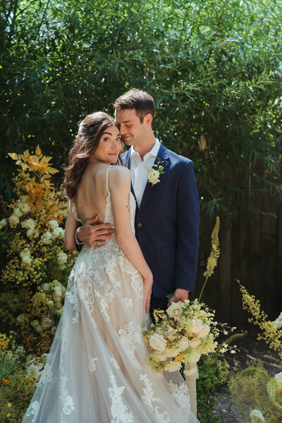 Bride in lacy, low-back wedding dress stands facing groom wearing navy blue suit stand outside at Frankie's 457 in Brooklyn surrounded by arranged spring flowers.