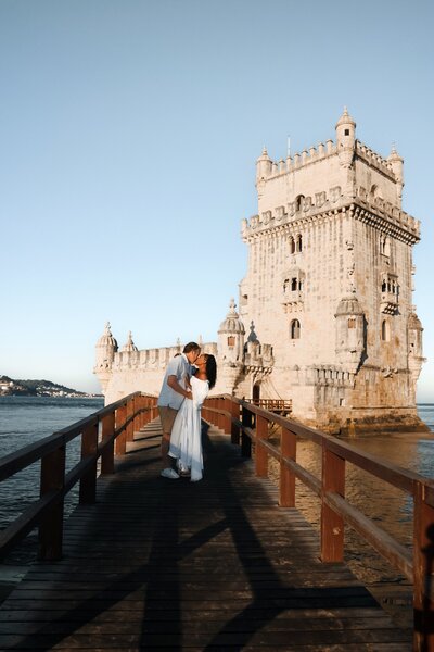 Couple embraces on a bridge leading to a tower in Lisbon