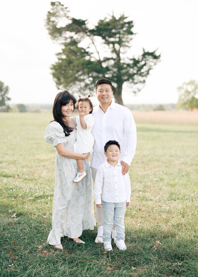 Woman wearing black floral dress and man wearing white button up shirt hold their daughter on a beach with trees in the background.