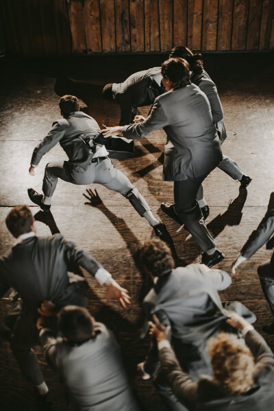 A group of groomsmen in gray suits are captured mid-motion on the dance floor, moving energetically in a choreographed or playful moment during the wedding reception.