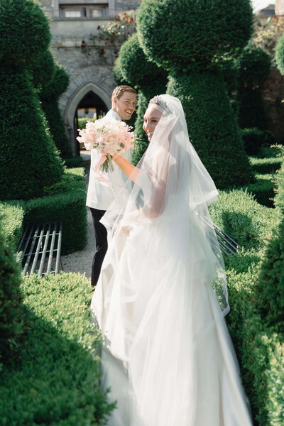 bride holding up her bouquet walking through euridge manor gardens with the groom and looking back at the camera