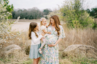 Expecting mom holds toddler and stands with young daughter laughing in field during maternity photoshoot