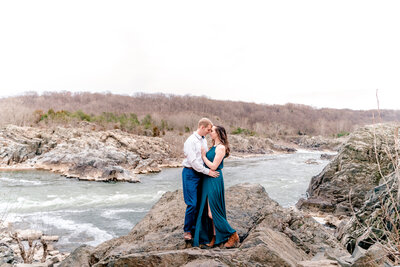 An engaged couple embrace while standing on the rocks above the Potomac River during their engagement session at Great Falls National Park