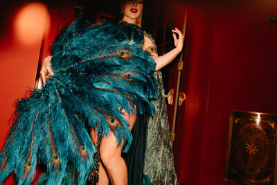 Close-up of a burlesque dancer holding a large teal feather fan with peacock eye patterns, wearing glamorous attire against a red backdrop.