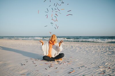 Older woman with white hair wearing a white long-sleeve shirt, jeans and an orange scarf sits cross-legged on the sand at the beach and throws colorful crayons in the air.