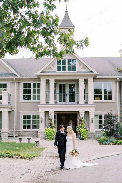 Bride and groom walking in front of Oakville Golf Club venue, holding hands and smiling, photographed by fine art Toronto wedding photographer