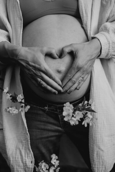 Schwarz weiss Bild von Babybauch mit Kirschblüten, Frau formt Herz mit ihren Händen vor ihrem Bauch