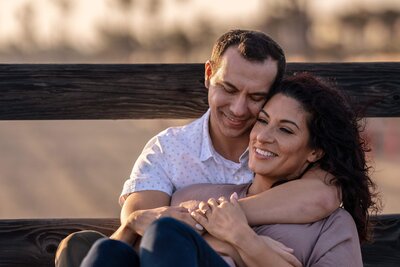 Loving couple embracing on an oceanside bench during sunset, captured in a fine art photography style, showcasing the intimate engagement session from Loma Linda photographer