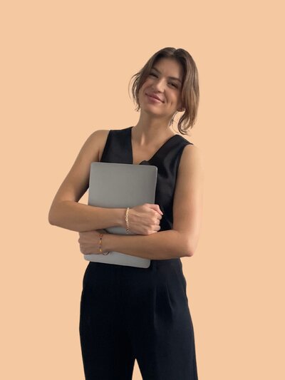 Caitlin holding a laptop close to her chest and smiling confidently. She wears a sleek black crop top and trousers against a soft yellow background.