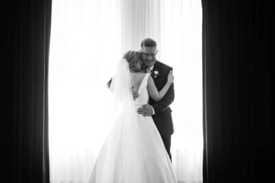 Black and White wedding photo of bride hugging her dad in front of a back lit window.