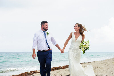 Laughing bride and groom holding hands walking on beach