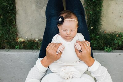 A baby sleeping in someone's lap, with adult hands holding the baby's hands, captured by a Pittsburgh photographer specializing in newborn lifestyle photography.